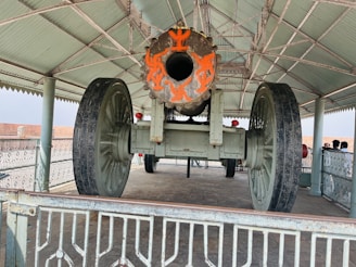 A large historical cannon is displayed under a metal canopy. The cannon has ornate orange markings on its barrel and is mounted on large spoked wheels. The area is surrounded by a decorative metal railing, and several people are visible in the background, standing near the structure.
