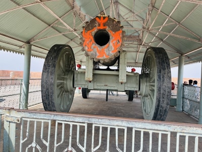 A large historical cannon is displayed under a metal canopy. The cannon has ornate orange markings on its barrel and is mounted on large spoked wheels. The area is surrounded by a decorative metal railing, and several people are visible in the background, standing near the structure.