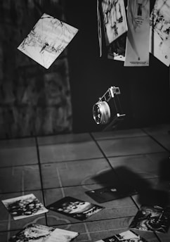 A vintage camera is suspended in mid-air along with several scattered photographs, some of which are blurred and others in focus, suggesting motion. The background has a dark, textured wall and a tiled floor is visible below.