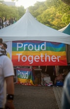 A tent is set up outdoors with a colorful banner that reads 'Proud Together'. The banner features rainbow stripes, commonly associated with the LGBTQ+ community. There are people walking around the area, and the surroundings include trees and a bright, sunny atmosphere.