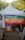 A tent is set up outdoors with a colorful banner that reads 'Proud Together'. The banner features rainbow stripes, commonly associated with the LGBTQ+ community. There are people walking around the area, and the surroundings include trees and a bright, sunny atmosphere.
