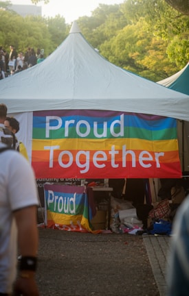 A tent is set up outdoors with a colorful banner that reads 'Proud Together'. The banner features rainbow stripes, commonly associated with the LGBTQ+ community. There are people walking around the area, and the surroundings include trees and a bright, sunny atmosphere.