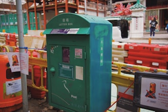 A green letter box stands on a city street surrounded by construction barriers and signs. The background shows construction work with orange and yellow barricades, signs in both English and Chinese, and a partially opened vehicle door. A few pedestrians are visible.