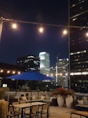 A rooftop patio at night featuring string lights and a blue patio umbrella. Comfortable seating areas are arranged around tables, and potted plants add a touch of greenery. The city skyline, with illuminated skyscrapers, forms a stunning backdrop.