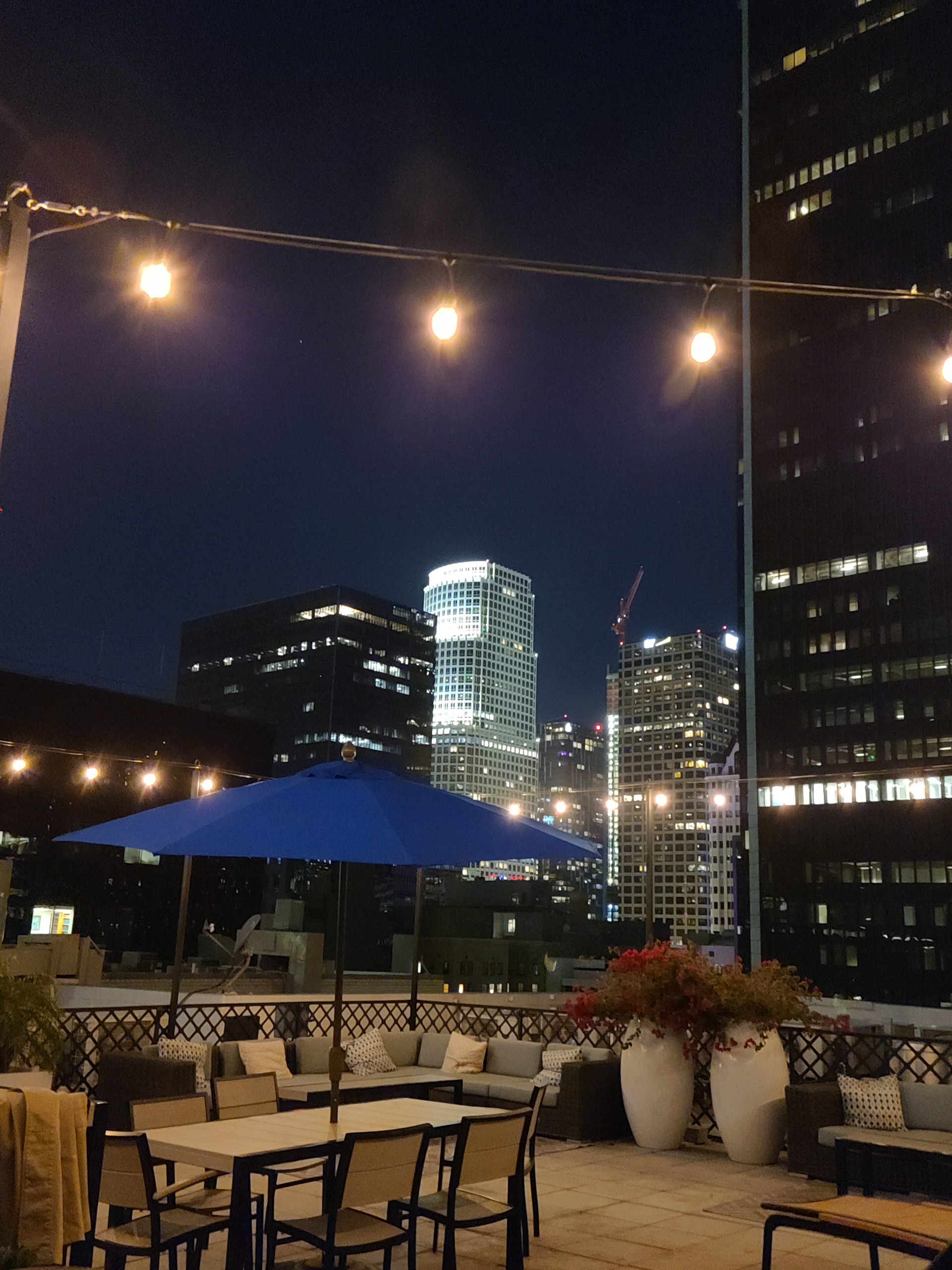 A rooftop patio at night featuring string lights and a blue patio umbrella. Comfortable seating areas are arranged around tables, and potted plants add a touch of greenery. The city skyline, with illuminated skyscrapers, forms a stunning backdrop.