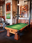 A well-furnished room with a pool table at the center. The pool table has a green felt surface and wooden legs. Hung above it are unique triangular pool table lights. The walls are adorned with various vibrant artworks and signs, including a green sign that reads 'RHODIAN BRANDS' and colorful paintings. There are wooden chairs and tables along the walls, creating a cozy seating area. The overall decor of the room is vintage with exposed brick walls and industrial elements.