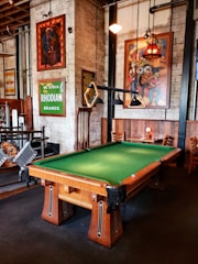 A well-furnished room with a pool table at the center. The pool table has a green felt surface and wooden legs. Hung above it are unique triangular pool table lights. The walls are adorned with various vibrant artworks and signs, including a green sign that reads 'RHODIAN BRANDS' and colorful paintings. There are wooden chairs and tables along the walls, creating a cozy seating area. The overall decor of the room is vintage with exposed brick walls and industrial elements.