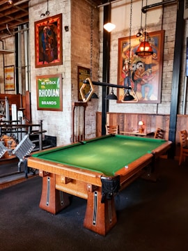 A well-furnished room with a pool table at the center. The pool table has a green felt surface and wooden legs. Hung above it are unique triangular pool table lights. The walls are adorned with various vibrant artworks and signs, including a green sign that reads 'RHODIAN BRANDS' and colorful paintings. There are wooden chairs and tables along the walls, creating a cozy seating area. The overall decor of the room is vintage with exposed brick walls and industrial elements.