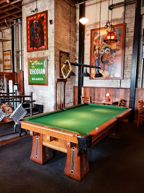 A well-furnished room with a pool table at the center. The pool table has a green felt surface and wooden legs. Hung above it are unique triangular pool table lights. The walls are adorned with various vibrant artworks and signs, including a green sign that reads 'RHODIAN BRANDS' and colorful paintings. There are wooden chairs and tables along the walls, creating a cozy seating area. The overall decor of the room is vintage with exposed brick walls and industrial elements.