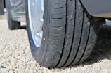 A close-up view of a car tire resting on a gravel-covered ground. The tire has a clean, well-defined tread pattern.