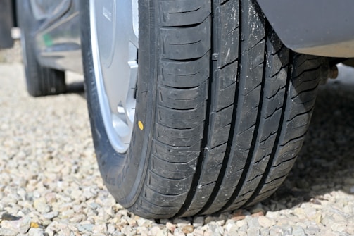 A close-up view of a car tire resting on a gravel-covered ground. The tire has a clean, well-defined tread pattern.