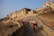 A long line of decorated elephants ascends a stone pathway leading up to a large, historic fort with intricate architecture and multiple levels. The structure is made of yellow sandstone and features traditional Rajasthani design elements including decorative windows and towers.