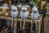 Three decorative bird figures with fluffy white heads and grey speckled bodies are sitting on a wooden shelf. They have long, rope-like legs with small metal feet. Behind them are other similar bird figures, featuring light brown beaks and cheerful expressions.