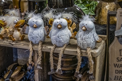 Three decorative bird figures with fluffy white heads and grey speckled bodies are sitting on a wooden shelf. They have long, rope-like legs with small metal feet. Behind them are other similar bird figures, featuring light brown beaks and cheerful expressions.