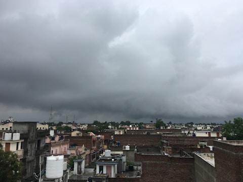 A densely populated urban area features an array of flat-roofed buildings, with various water tanks visible on top. The sky is heavily overcast with thick dark clouds, indicating an impending storm. Buildings vary in color from beige to pink, and the scene includes some greenery in the distance.