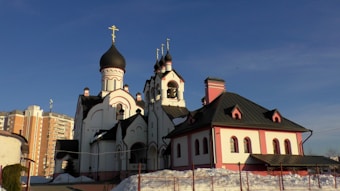 An Orthodox church featuring multiple domes with crosses on top, characterized by its white and pink exterior with dark roofing. The building exhibits traditional architectural elements such as arched windows and a geometric design. In the background, there are residential buildings. Snow is visible on the ground, indicating a winter setting.