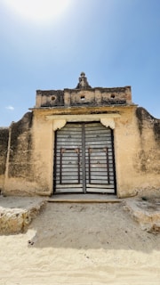 A weathered stone manor gate with iron details, framed by ancient trees under a soft, cloudy sky.
