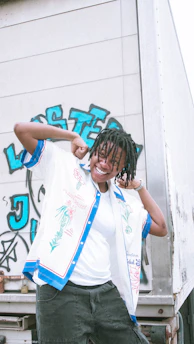 A person with dreadlocks is smiling and posing energetically in front of a truck with graffiti art on its side. They are wearing a colorful shirt with floral designs over a white t-shirt and dark pants.