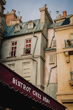 A charming building with typical Parisian architecture, featuring round windows, chimneys, and a variety of textures and colors in the facade. The structure appears aged and has elements like red shutters and green roofing. Below, an awning displays the name 'Bistrot Chez Rémy' in white letters on a burgundy background.