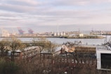 A river or harbor area is visible with industrial structures such as cranes, wind turbines, and storage tanks. In the foreground, there are several rows of brick buildings partly obscured by trees, with a large bridge in the background crossing over the water.
