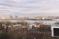 A river or harbor area is visible with industrial structures such as cranes, wind turbines, and storage tanks. In the foreground, there are several rows of brick buildings partly obscured by trees, with a large bridge in the background crossing over the water.