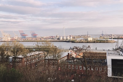 A river or harbor area is visible with industrial structures such as cranes, wind turbines, and storage tanks. In the foreground, there are several rows of brick buildings partly obscured by trees, with a large bridge in the background crossing over the water.