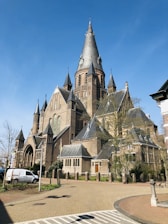 A grand historic church with tall spires and intricate architectural details made of brick and stone, set against a clear blue sky. The building is surrounded by small trees and a paved pathway, with a white van parked nearby.