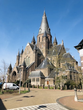 A grand historic church with tall spires and intricate architectural details made of brick and stone, set against a clear blue sky. The building is surrounded by small trees and a paved pathway, with a white van parked nearby.