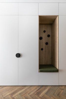 A minimalist built-in cabinet with a white exterior and a recessed wooden alcove. The alcove has a small green cushion and six black pegs arranged on the back wall. The floor features a herringbone wood pattern.