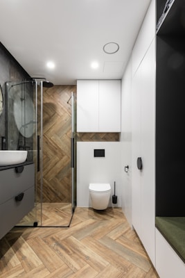 A modern bathroom featuring wooden herringbone-patterned flooring and walls. A sleek glass shower enclosure with black fixtures sits next to a wall-mounted toilet. Minimalistic white cabinets offer storage. The room is well-lit with recessed ceiling lights.