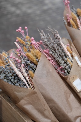 Bouquets of dried flowers wrapped in brown paper, featuring a variety of textures and colors. The flowers include pink buds, yellow grasses, and muted blue and brown botanicals.