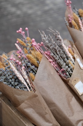 Bouquets of dried flowers wrapped in brown paper, featuring a variety of textures and colors. The flowers include pink buds, yellow grasses, and muted blue and brown botanicals.