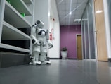 A white humanoid robot is positioned in a hallway of an office or building. The corridor has a tiled floor and is lined with shelving on one side and doors on the other. The back wall is painted purple and features a potted plant. Overhead, fluorescent lights illuminate the space, and a fire extinguisher is mounted on the wall.