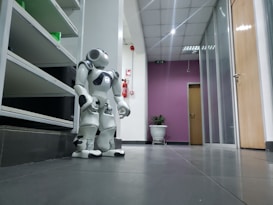 A white humanoid robot is positioned in a hallway of an office or building. The corridor has a tiled floor and is lined with shelving on one side and doors on the other. The back wall is painted purple and features a potted plant. Overhead, fluorescent lights illuminate the space, and a fire extinguisher is mounted on the wall.