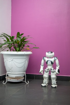 A humanoid robot stands on the floor next to a large white plant pot with green leaves. The background features a bright pink wall, creating a contrast with the grey floor tiles.