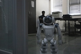 A humanoid robot stands on a tiled floor in an office setting. Behind the robot, a person is seated at a desk, partially illuminated by light coming through window blinds. The office atmosphere includes furniture like a cabinet, desk, and office equipment such as a printer.
