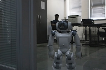 A humanoid robot stands on a tiled floor in an office setting. Behind the robot, a person is seated at a desk, partially illuminated by light coming through window blinds. The office atmosphere includes furniture like a cabinet, desk, and office equipment such as a printer.