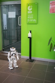 A humanoid robot stands on the tiled floor near a bright green wall that displays a sign for the Goethe-Institut along with its opening hours. A sanitizer dispenser is mounted on a stand near the robot.