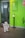 A humanoid robot stands on the tiled floor near a bright green wall that displays a sign for the Goethe-Institut along with its opening hours. A sanitizer dispenser is mounted on a stand near the robot.