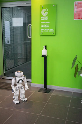 A humanoid robot stands on the tiled floor near a bright green wall that displays a sign for the Goethe-Institut along with its opening hours. A sanitizer dispenser is mounted on a stand near the robot.