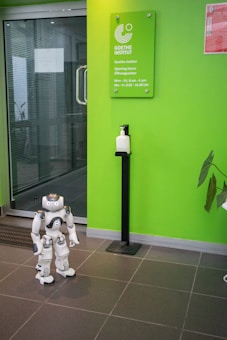 A humanoid robot stands on the tiled floor near a bright green wall that displays a sign for the Goethe-Institut along with its opening hours. A sanitizer dispenser is mounted on a stand near the robot.