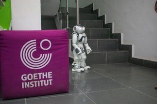 A small humanoid robot stands at the base of a staircase with tiled steps. To the left, a purple cube with the logo and words 'Goethe Institut' is visible. The floor is also tiled, and the surrounding walls are mostly plain.