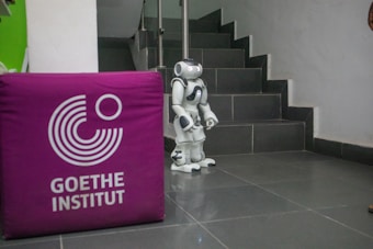 A small humanoid robot stands at the base of a staircase with tiled steps. To the left, a purple cube with the logo and words 'Goethe Institut' is visible. The floor is also tiled, and the surrounding walls are mostly plain.