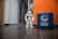 A humanoid robot stands on a shiny tiled floor in a hallway next to a blue cube with the text 'Goethe Institut' printed on it. The background features a glass door and walls in white and orange colors.