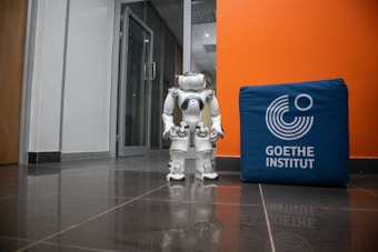 A humanoid robot stands on a shiny tiled floor in a hallway next to a blue cube with the text 'Goethe Institut' printed on it. The background features a glass door and walls in white and orange colors.