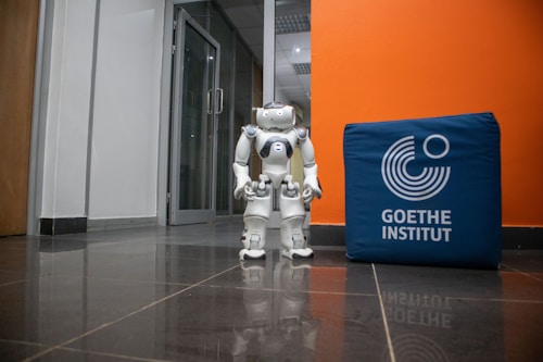 A humanoid robot stands on a shiny tiled floor in a hallway next to a blue cube with the text 'Goethe Institut' printed on it. The background features a glass door and walls in white and orange colors.