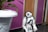 A small humanoid robot with a white and black exterior stands in front of a wooden door. There is a large white plant pot with green leaves in the foreground. The wall behind the robot is painted in a vibrant purple color.