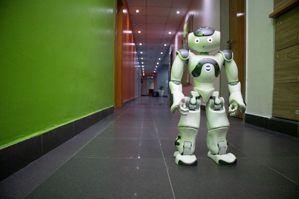 A humanoid robot stands in a corridor with a green wall on one side and wooden paneling on the other. The robot is predominantly white with black accents and appears to have articulated joints. The corridor is well-lit, with a ceiling lined with recessed lights.