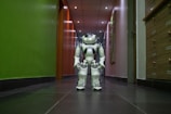 A humanoid robot with white and gray accents stands in the middle of a hallway. The environment features a dark floor and a brightly lit ceiling with recessed lights. The walls are painted green on one side and light on the other, with various wooden panels and doors visible.