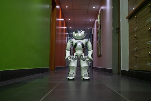 A humanoid robot with white and gray accents stands in the middle of a hallway. The environment features a dark floor and a brightly lit ceiling with recessed lights. The walls are painted green on one side and light on the other, with various wooden panels and doors visible.